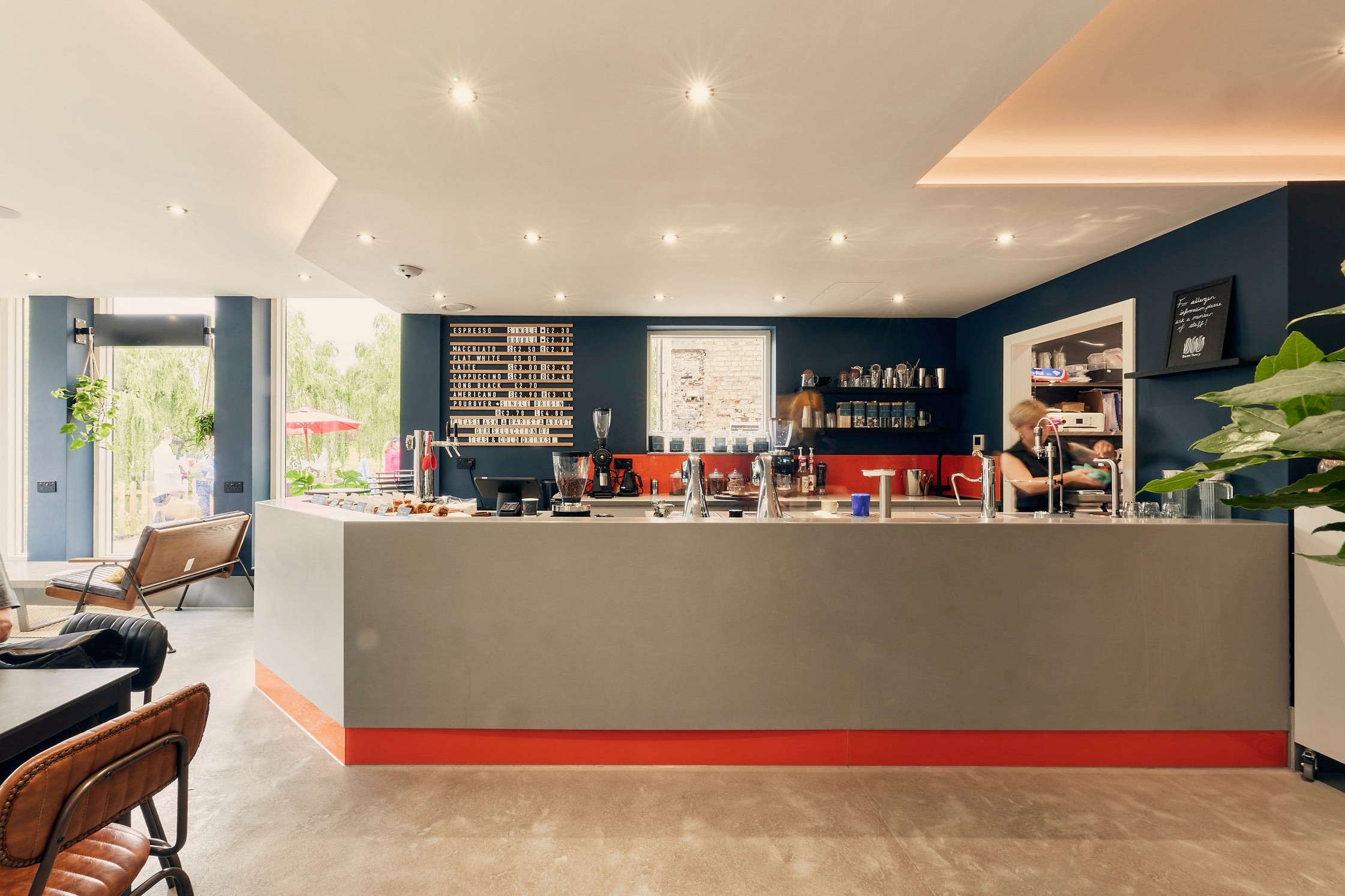 The counter at Bean Theory in Newnham, with a hand lettered coffee menu board, exposed brick, a red accent stripe along the base of the counter and a view through large windows to outdoor seating by the Mill Pond