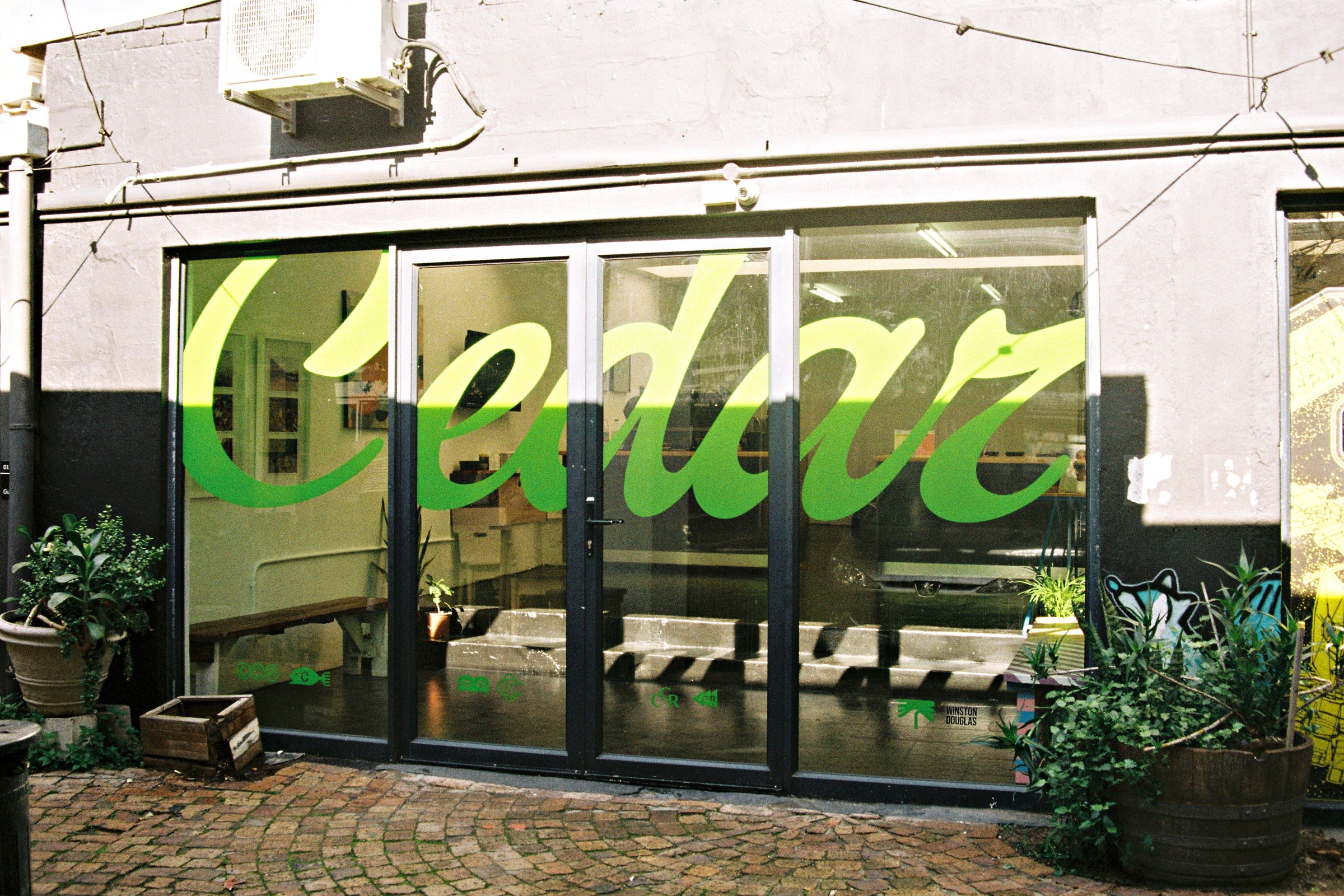 The glass shopfront at Side Street Studios in Woodstock with large green script Cedar lettering across the windows and doors, potted plants and a wooden crate on a cobbled lane