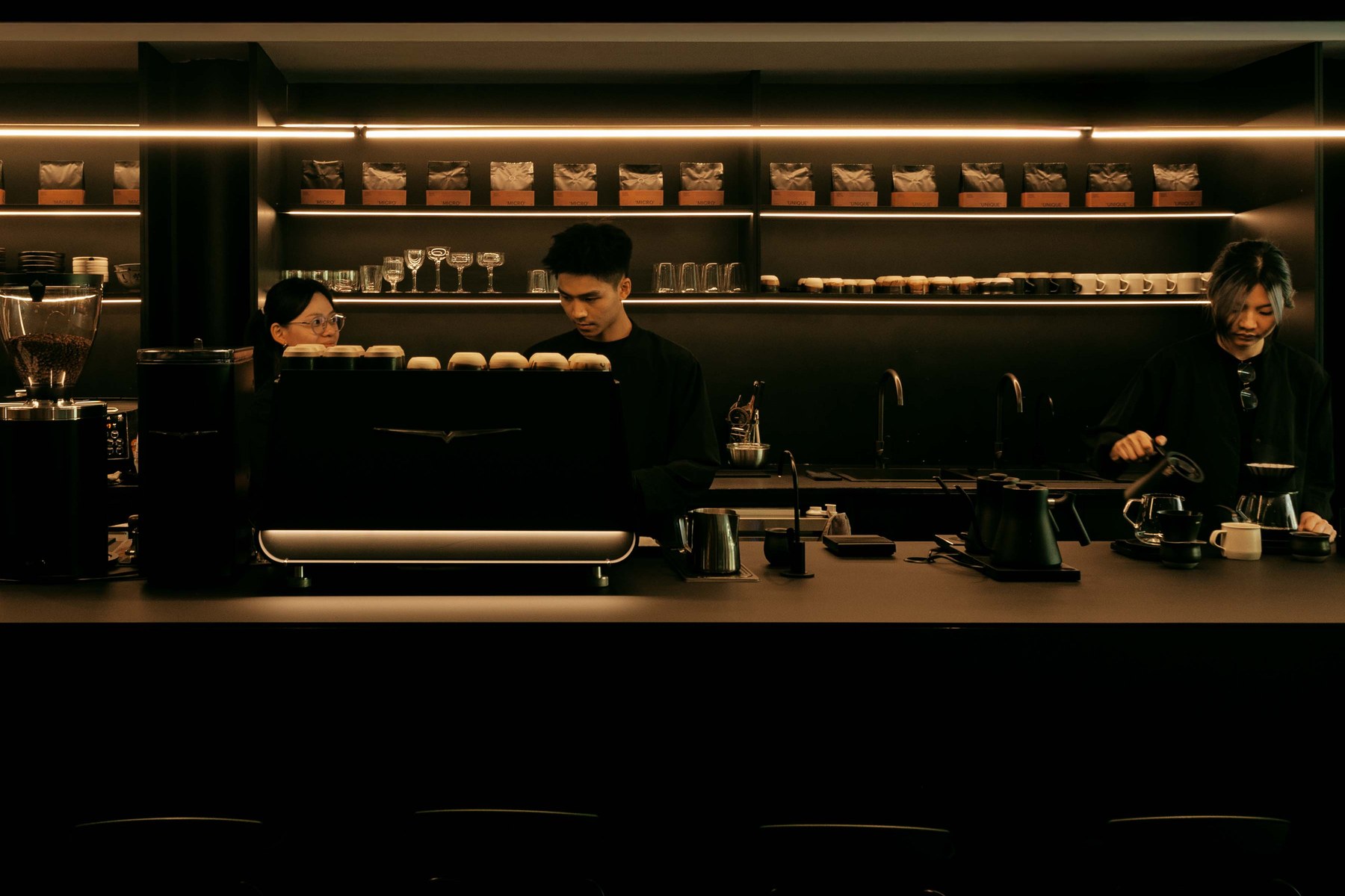The long black bar at Afloat Coffee Roaster with an espresso machine, shelves of bagged beans glowing under warm strip lights and baristas working behind the counter