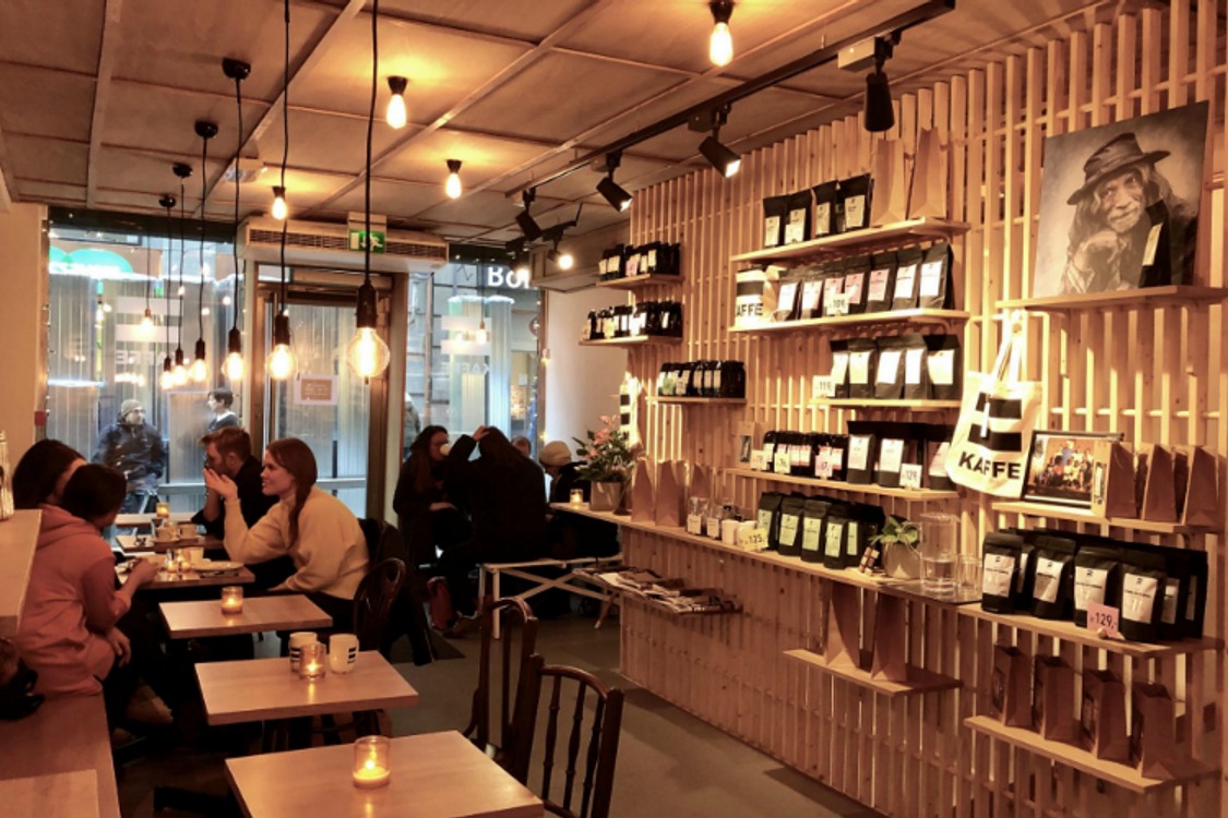The warm wood shelved interior of a Kaffe coffee bar, lined with bagged beans and a KAFFE tote bag, with customers seated at small tables