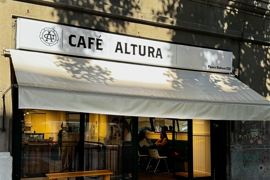 The storefront of Café Altura at Paseo Bulnes 122 with its white sign, a pale awning, a wooden bench out front and a warmly lit dining room behind the glass
