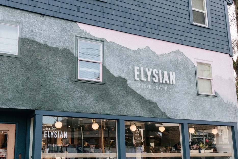 The corner storefront of Elysian Coffee at Broadway and Ash, with a hand painted mountain mural across the stucco wall, the Elysian Coffee Roasters name in white letters and warm pendant lights glowing in the windows
